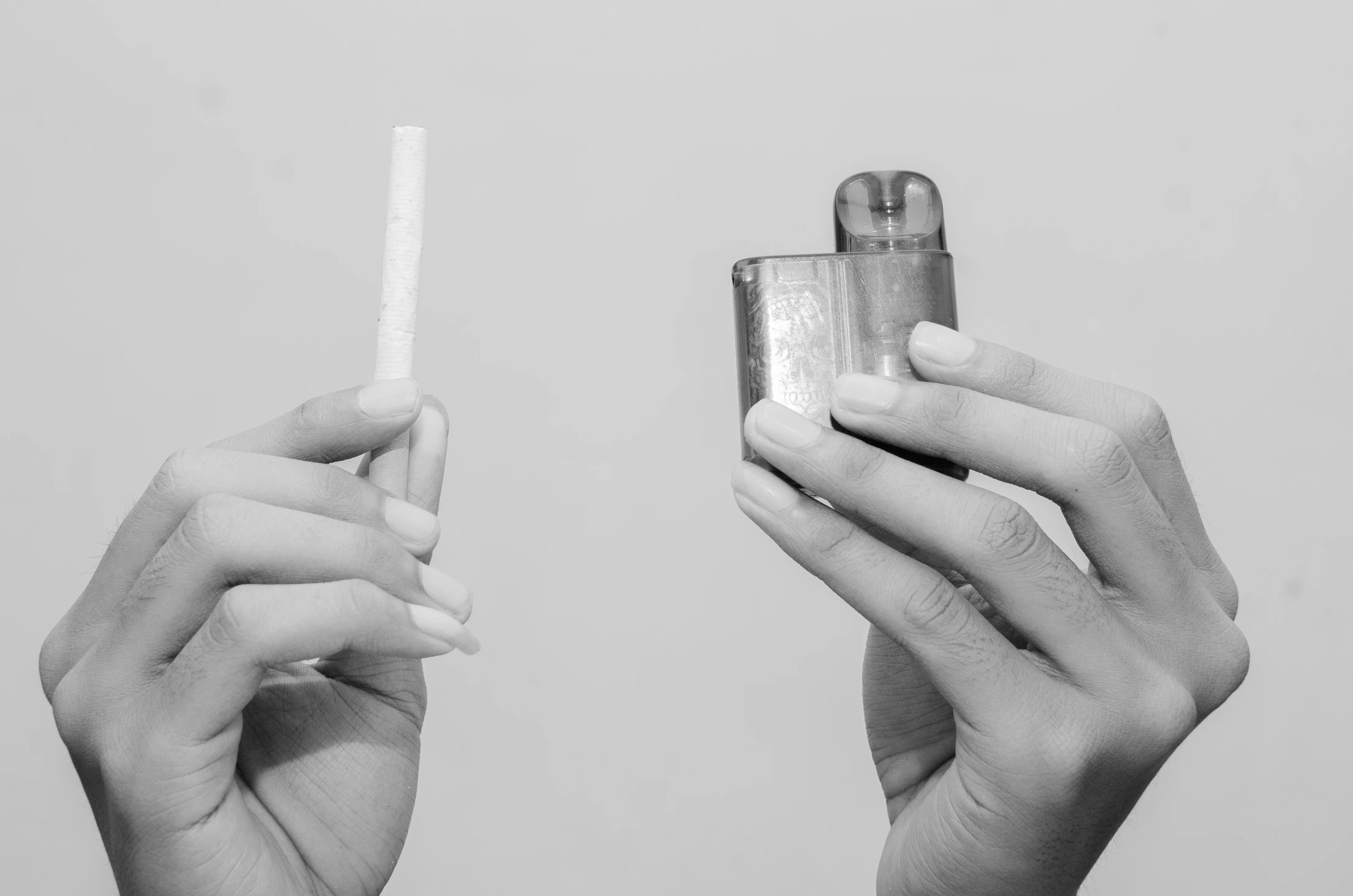 Black and white image of hands holding a vape kit and a cigarette for comparison