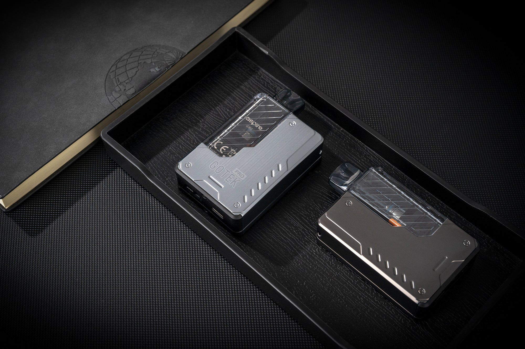 Two metallic Gotek Pro Vape devices on a dark textured surface with a logo in the background